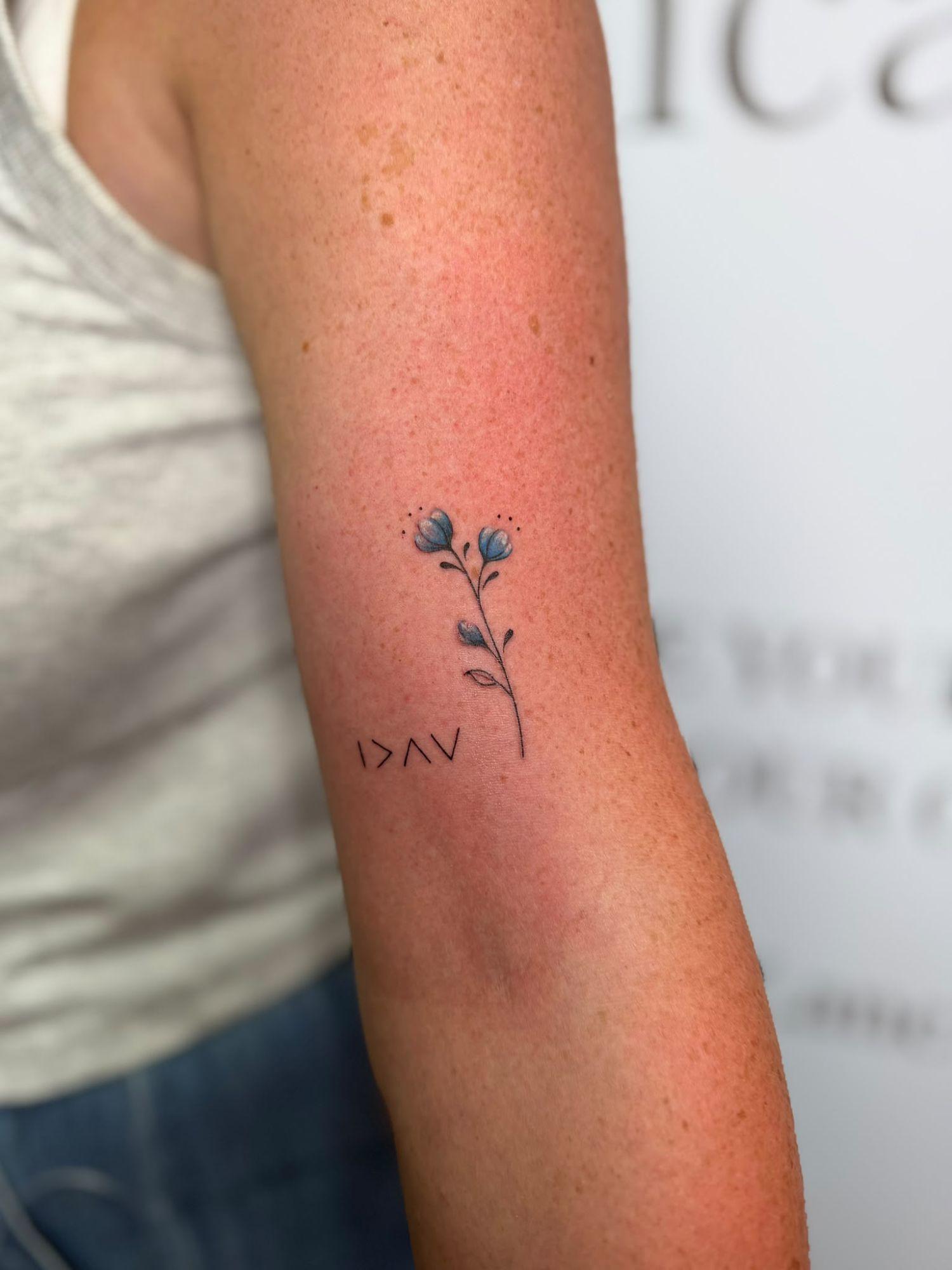 Small blue flower tattoo with a fine-line symbol on an upper arm